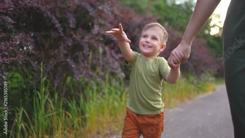 A woman and a boy walk together on a path surrounded by greenery and purple bushes. The boy smiles and holds the woman's hand as they enjoy the day.