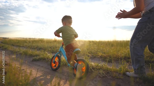 A woman encouraging a boy riding a blue bike in a grassy field. The boy pedals his bike while the woman applauds his efforts.