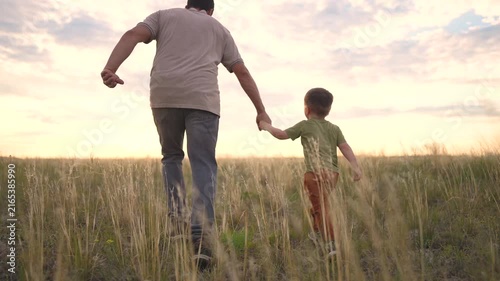 A man and a boy walk together in a field, holding hands. The man and the boy enjoy the view as they walk through the tall grass.