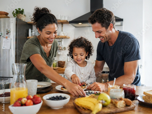 A mother and father help a curly haired toddler arrange fruit and toast on a kitchen counter loaded with berries, juice and bread during a bright and cheerful family breakfast.