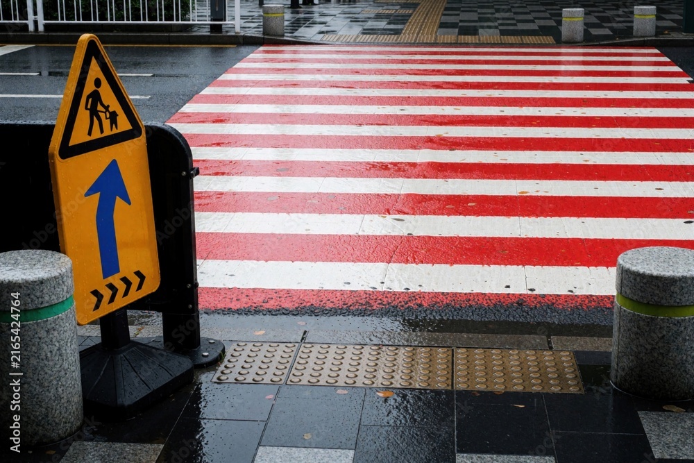Fototapeta premium Bright red, white pedestrian crosswalk on wet asphalt road. Yellow traffic warning sign shows pedestrian crossing direction. Urban street view after rain with colorful markings, safety infrastructure