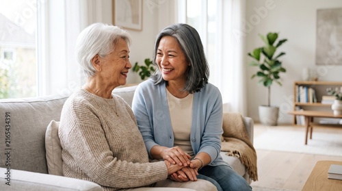 Two women, senior and middle-aged, sit on a sofa holding hands and smiling at each other in a bright home living room, conveying family love, support, and care