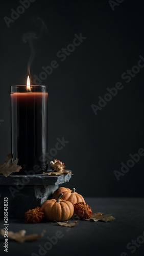 A tall black candle in a glass holder with smoke, small pumpkins, and dried autumn leaves on a dark stone surface against a dark background.