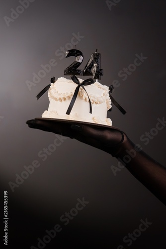Hand in black glove holding small white cake with black ribbons and number 24 candles on dark grey background.