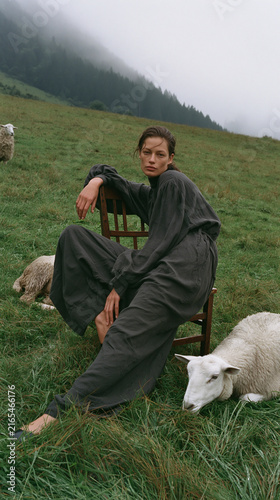 Moody Rural Fashion Portrait of Woman Sitting Among Sheep in a Misty Mountain Meadow