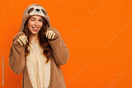 Playful woman in sloth costume winks and sticks out tongue against Halloween orange background with copy space