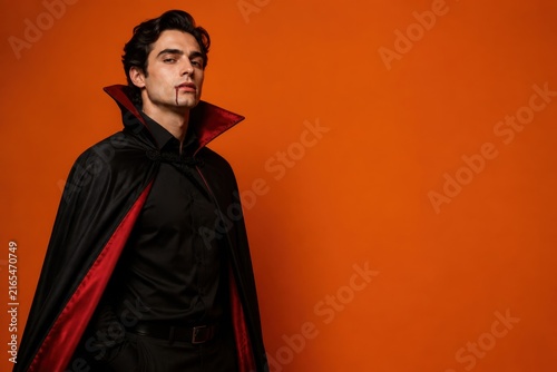 Man dressed as a vampire with blood dripping from his mouth against Halloween orange background with copy space