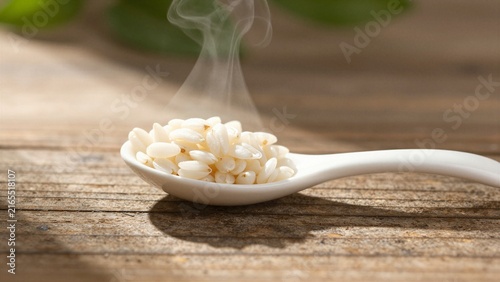 A white ceramic spoon filled with steaming uncooked rice grains rests on a rustic wooden table.
