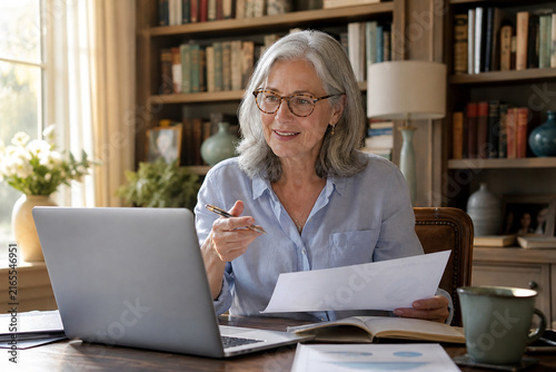 Experienced Senior Female Consultant Reviewing Business Documents during a Video Call from an Elegant Home Study with Warm Bookshelves