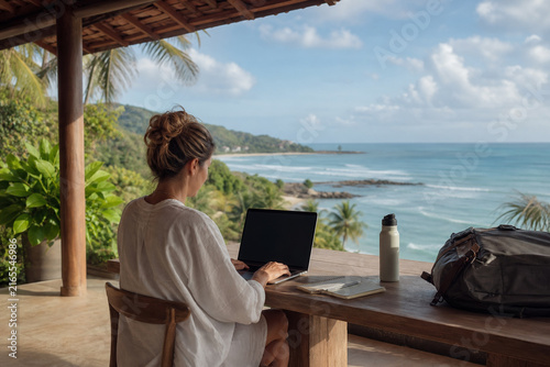 Female Digital Nomad Working on a Laptop from a Shaded Tropical Terrace overlooking a Peaceful Ocean Coastline during a Flexible Workday