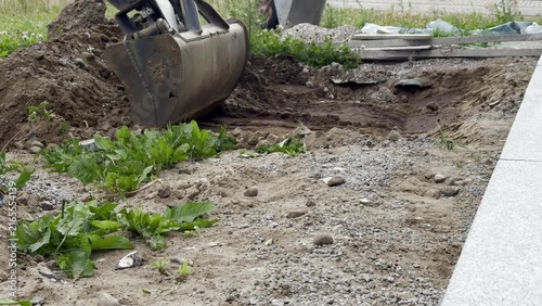 Excavator bucket hovers over a dirt mound at a construction site, with grass and gravel in the foreground 