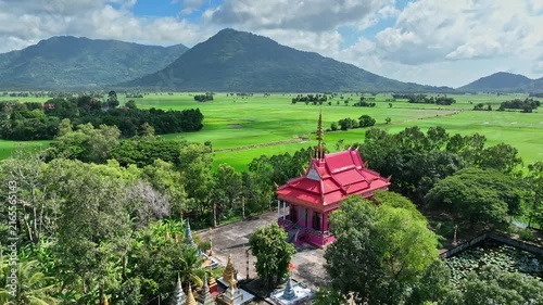 Aerial landscape of a vivid red Buddhist temple surrounded by trees, rice fields, and lush mountains in An Giang Province, Vietnam.
