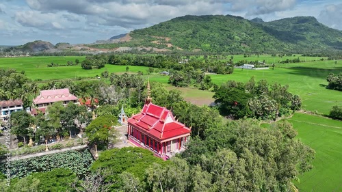 Drone view of a vibrant Buddhist temple surrounded by lush rice fields, tropical countryside, and green mountains in An Giang, Vietnam.
