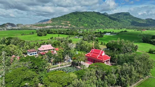 Aerial view of a colorful Buddhist pagoda surrounded by lush trees, rice fields, rural homes, and green mountains in An Giang, Vietnam.