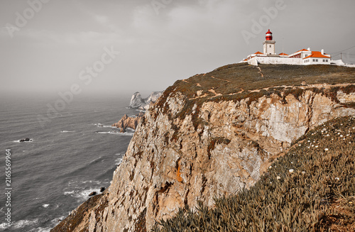 Lighthouse at Cape Roca (Cabo da Roca), most western point of Europe at coast of Atlantic Ocean in Portugal. Rocks at coastline and blue sky with clouds over skyline. Picturesque summer landscape.
