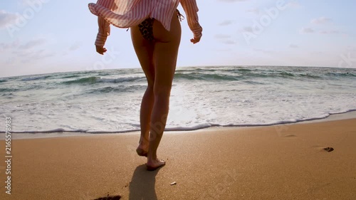 The young slender woman in a stylish swimsuit and shirt walks on the sandy beach, looking at the calm ocean horizon with a sense of freedom and tranquility, travel and summer lifestyle.