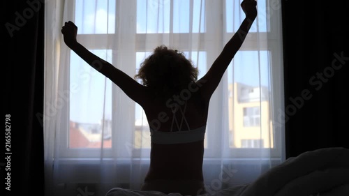 Lovely woman with curly hair stretching her arms after waking up, sitting on a bed in a dark room. Silhouette of her body against a bright window starting a beautiful new day at home