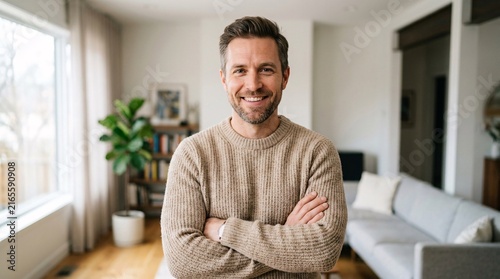 Confident man in a cozy beige knit sweater with arms folded, smiling warmly at the camera in a bright airy living room with crisp detail.