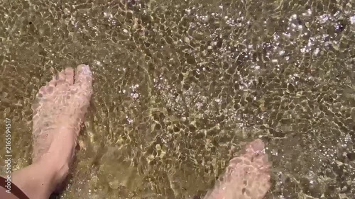 Close up first person view of bare feet touched by gentle sea waves on warm sand creating a relaxing summer beach travel and wellness concept. High quality FullHD footage
