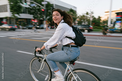 Smiling young woman with a backpack rides a bicycle on a busy urban street with motion blur, looking back at the camera, embodying an active, healthy, and eco-friendly city lifestyle during