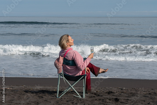 Foto Woman sits in folding chair holding glass of champagne beside Pacific Ocean on black volcanic beach in Kamchatka