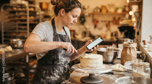 Professional pastry chef decorating layer cake while following digital recipe on mobile phone screen.