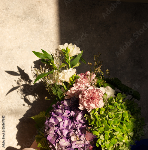 Floral arrangement featuring purple hydrangea, carnation, and freesia flowers in a vase illuminated by direct sunlight, creating dramatic hard light shadows on a neutral stone background.