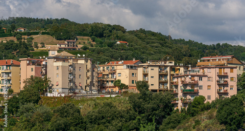 Vista panorámica de un pueblo mediterráneo con coloridos edificios residenciales en la ladera de una colina verde bajo un cielo nublado.
