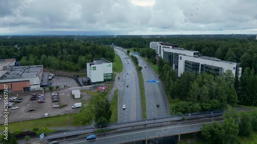 Cinematic aerial drone footage over Pohjois-Haaga, Helsinki, Finland, featuring the Veikkaus building, surrounded by lush green summer landscapes under a cloudy sky.