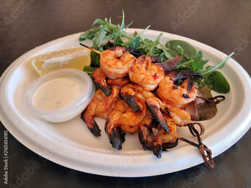Foto Grilled shrimp skewers served with fresh arugula, baby spinach, a lemon wedge, and white sauce dip on a paper plate