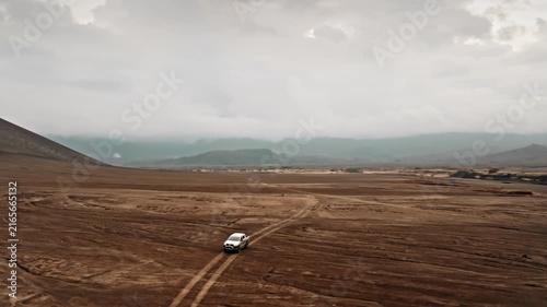 White truck driving on dirt track across the vast, rugged volcanic ash plain with Mount Yasur volcano slope looming under a cloudy sky in Tanna Island, Port Resolution. Aerial view drone panorama