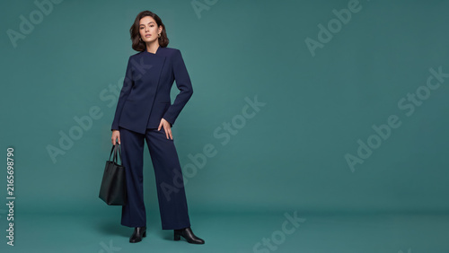 A confident woman models a navy suit and boots. She holds a leather tote bag while posing in a teal studio, leaving an empty area for text.