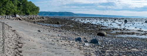 Fotografie Panoramic view of a foreshore exposed at low tide, characterized by piles of rocks, seaweed and pebbles with a view of the mountains along the St