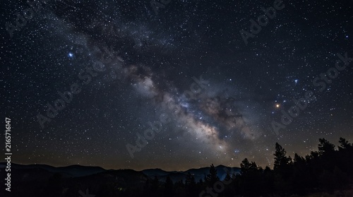 Starry night sky with Milky Way galaxy over dark forested mountain landscape at dusk