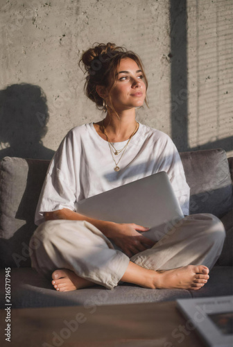 Young woman sitting cross-legged on sofa holding closed laptop in sunny room