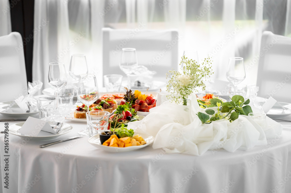 Fototapeta premium Elegant wedding table setting with white tablecloth, delicate flower centerpieces, wine glasses, and plates filled with gourmet appetizers and cheese platters in a brightly lit banquet hall.