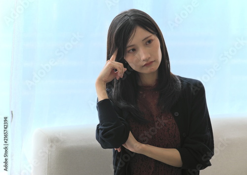 Фотография 窓際で頭に指をあてて深く悩み考える若い女性（悩み・思考・ストレスイメージ）