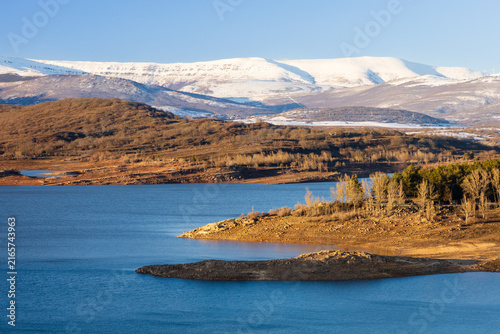 Scenic winter landscape of Aguilar Reservoir with rolling shorelines and snow-capped mountain peaks in the distance. Aguilar de Campoo, Palencia, Castile and Leon, Spain.