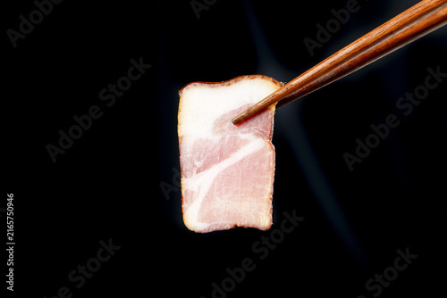 Konstfotografi Raw bacon slice held by wooden chopsticks on black background - meat cooking ing