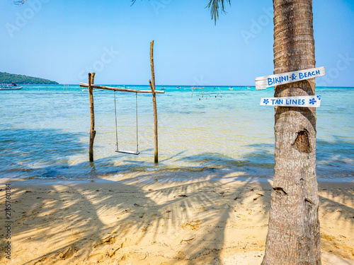 Alano Beach in Karimunjawa, Indonesia, featuring a wooden swing in shallow turquoise water next to a palm tree with Bikini and Beach and Tan Lines signs, creating a serene tropical travel destination
