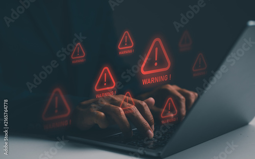 A person typing on a laptop surrounded by multiple red warning signs indicating cyber threats or security alerts.
