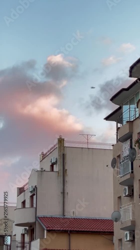 Soft, warm sunset light illuminates dramatic puffy clouds above residential apartment buildings with terracotta tile roofs and balconies.