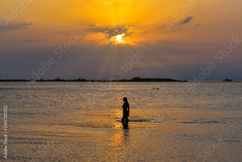 Silhouette of a person walking through the shallow sea at sunset in Glyfada, Athens Riviera, Greece, with golden sunlight reflecting across the water and dramatic clouds over the Saronic Gulf.
