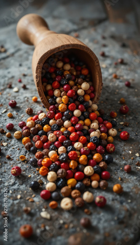 Wooden scoop spills colorful assortment of various peppercorns onto textured stone surface, creating vibrant and visually appealing display. mix includes shades of red, black, white, and brown