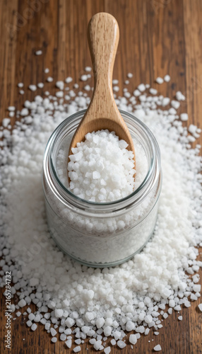 Glass jar filled with coarse salt sits wooden surface, surrounded by scattered salt grains. wooden spoon rests inside jar, emphasizing natural and rustic feel of scene. image evokes sense