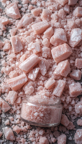 Natural pink salt crystals scattered around glass jar filled with fine pink salt. texture and color create calming and aesthetic appeal, perfect for culinary or spa use