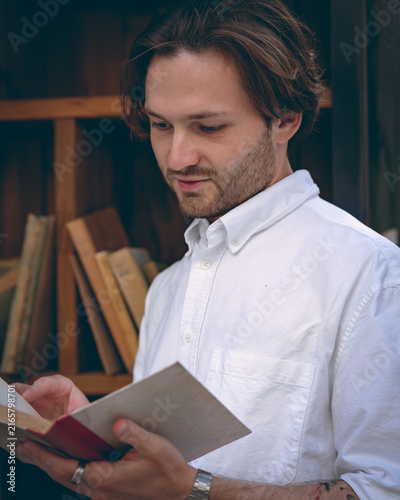 Fotografie Close-up of a young man in a white short-sleeved shirt turning a page while reading a book near a wooden bookshelf outdoors
