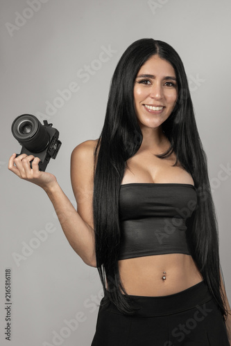 Beautiful young woman with straight black hair wearing a black top and outfit while holding a professional camera during a fashion studio photoshoot