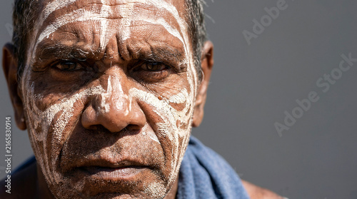 Close up portrait of an Australian Aboriginal elder with traditional white clay face paint, showcasing deep wrinkles and an intense spiritual gaze toward the camera against a neutral background.
