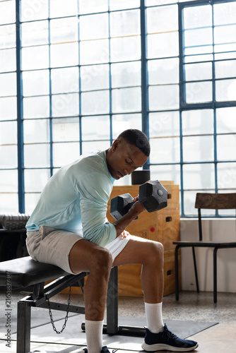 African man performing seated hex dumbbell curl on bench at gym, wearing light blue top
